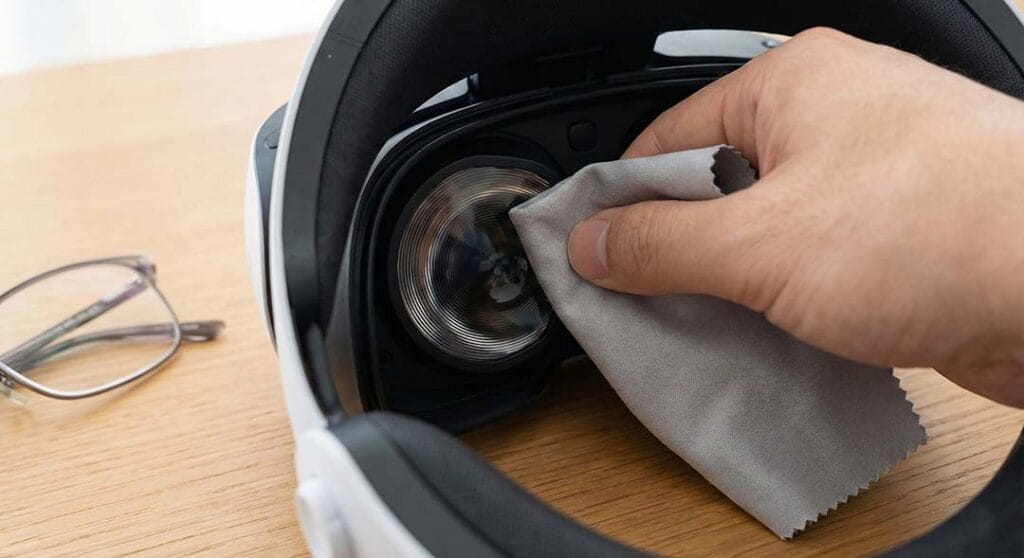 Hand gently cleaning PS VR2 internal lenses with a microfiber cloth next to eyeglasses.