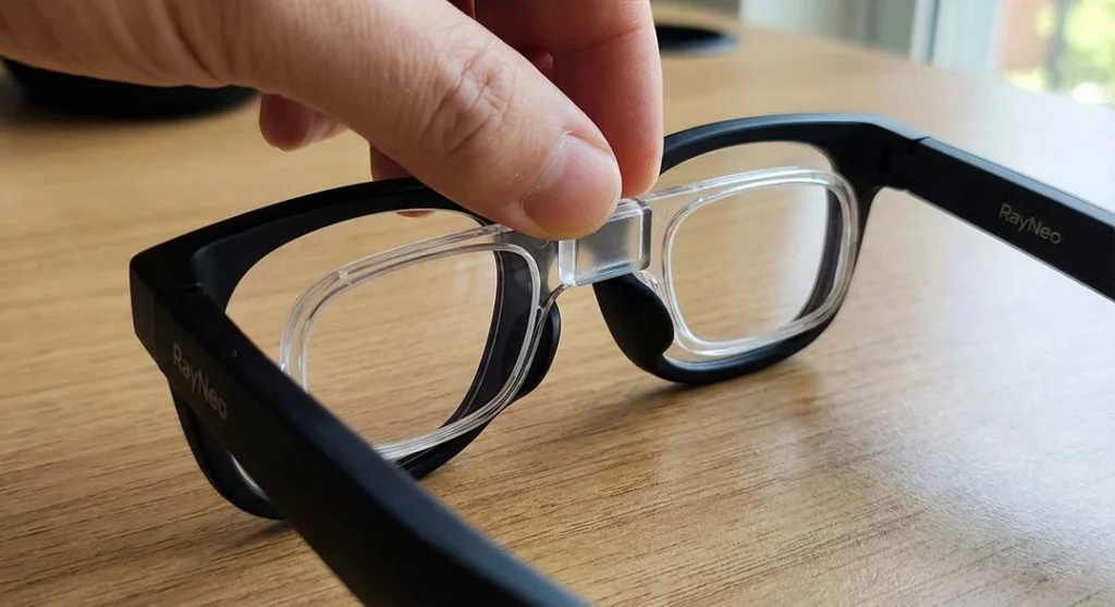 Close-up view of a hand snapping the ANNKUTVR prescription lens insert onto the nose bridge mechanism of RayNeo Air 2s AR glasses.